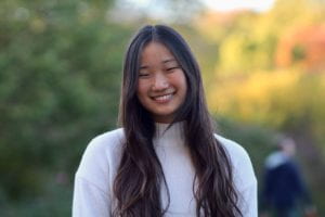 A picture of student Abigail Chung. She is wearing a white sweater and smiling at the camera in front of a natural background.